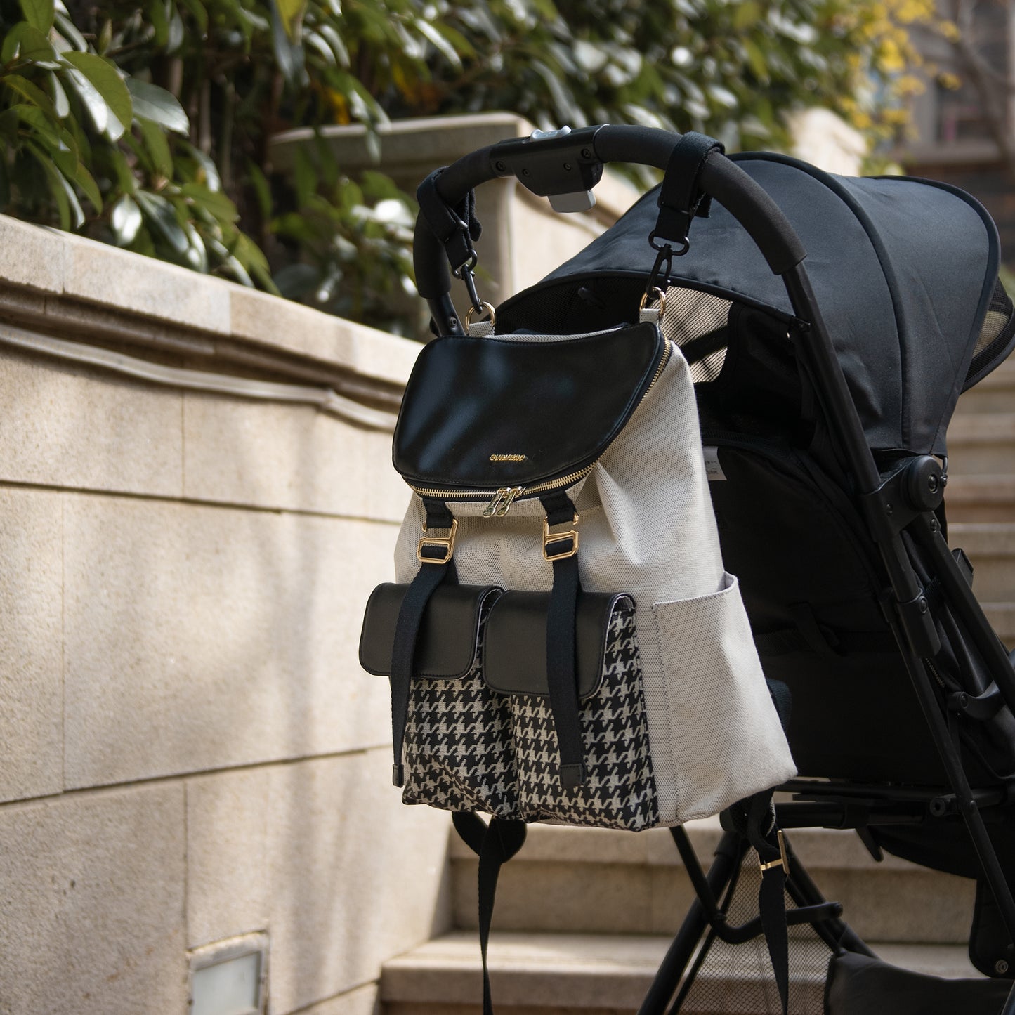 Black backpack with gold accents attached to a stroller against a stone wall background