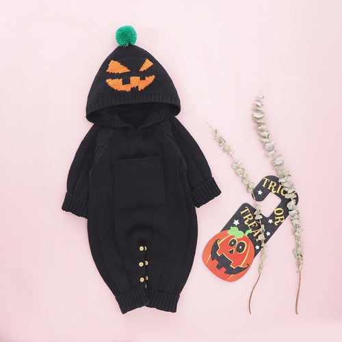 An orange baby romper with a monster print, including a hood with a spooky face design, displayed next to a small decorative pumpkin and a Halloween-themed pin.