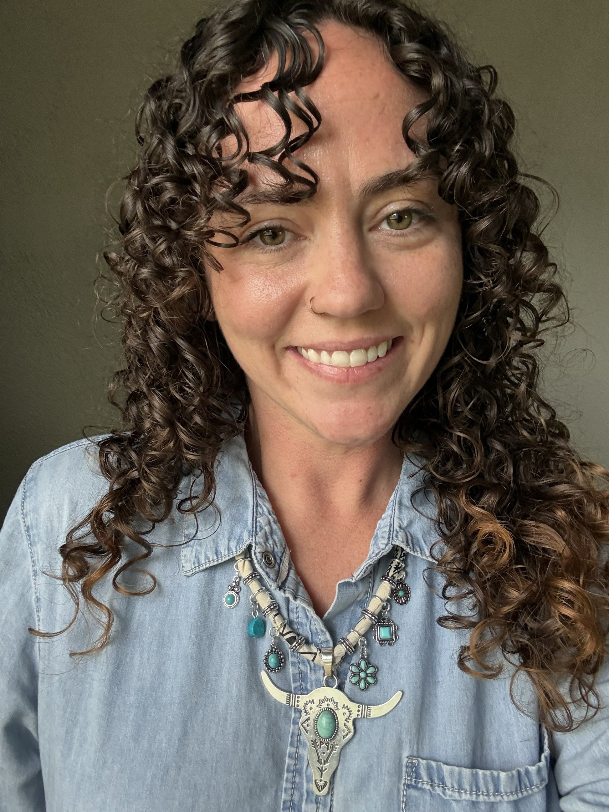 Woman with curly hair wearing a blue denim shirt and decorative necklace against a neutral background