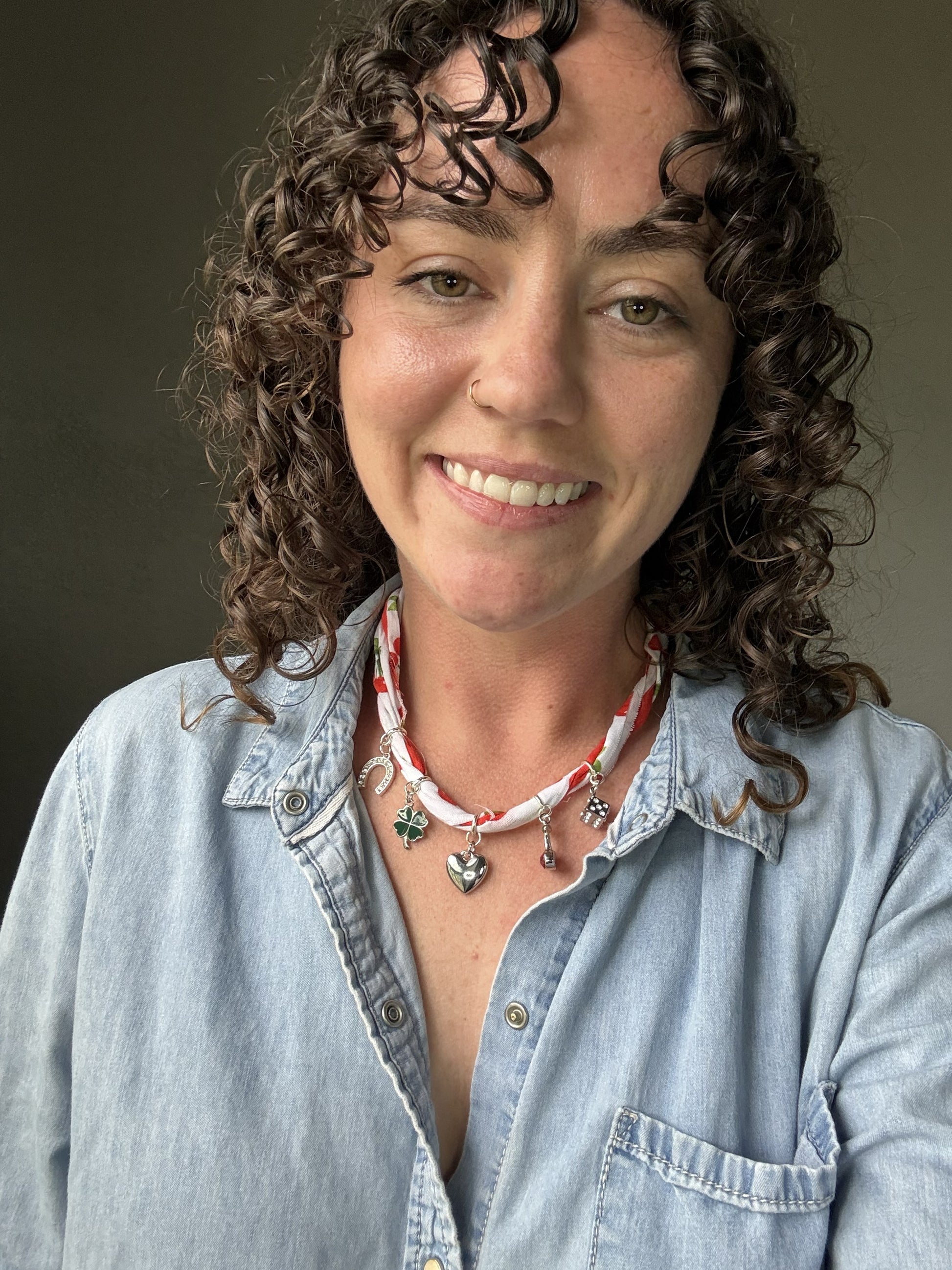 Woman with curly hair wearing a denim shirt and multiple necklaces against a neutral background