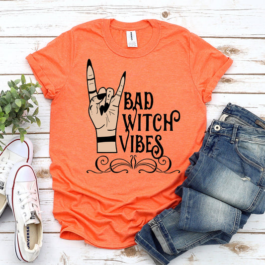 Orange t-shirt with 'Bad Witch Vibes' design, paired with jeans and sneakers on a wooden surface.