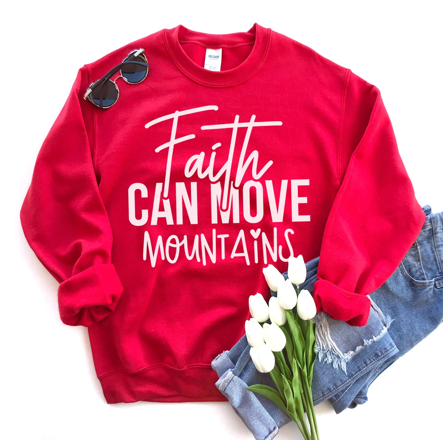 Red sweatshirt with 'Faith Can Move Mountains' text, sunglasses, and white tulips on a white background