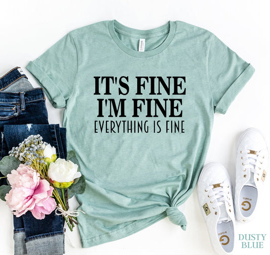 Mint green t-shirt with 'It's Fine, I'm Fine, Everything is Fine' text, paired with jeans, flowers, and sneakers on a white background.