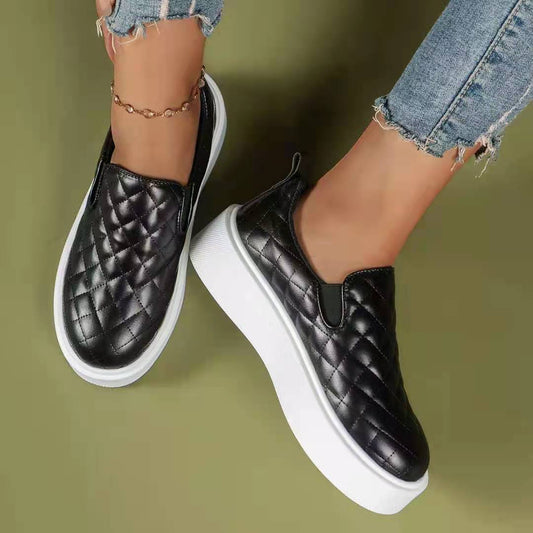 A pair of black slip-on casual platform shoes with a quilted pattern and thick platform sole, worn by a person with jeans.