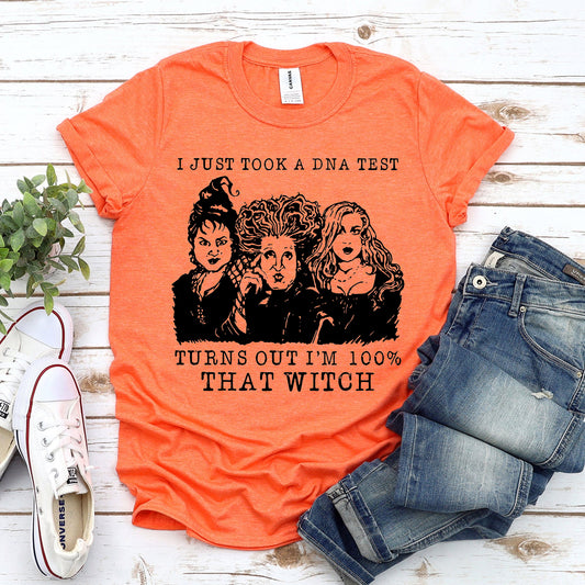 Orange t-shirt with text and graphic print depicting three witch characters, paired with jeans and white sneakers, displayed on a wooden surface.