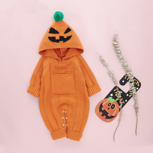 An orange baby romper with a monster print, including a hood with a spooky face design, displayed next to a small decorative pumpkin and a Halloween-themed pin.